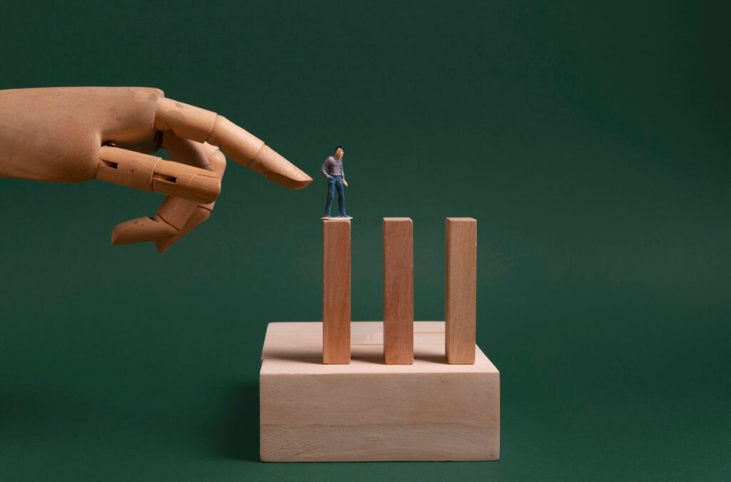 A wooden hand pointing at a small figurine standing on a wooden block, symbolizing the challenges of self-doubt and the importance of confidence in professional growth with Tadah!'s coaching support