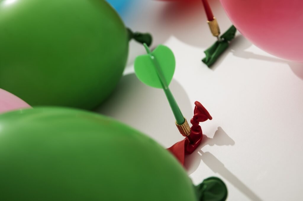 green dart piercing a deflated red balloon surrounded by green and pink balloons, symbolizing focus, precision, and overcoming challenges with Tadah!'s coaching strategies for professional growth and resilience